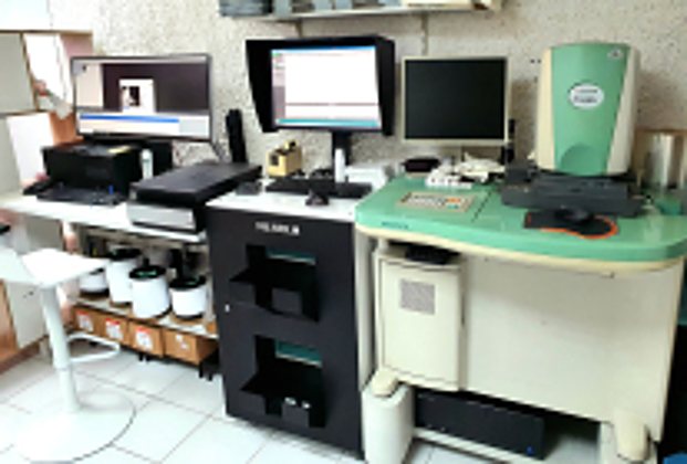 Poste de travail d'un labo photo avec écrans d'ordinateur, scanner et grande machine d'impression Fujifilm noire à côté d'un appareil vert et blanc.