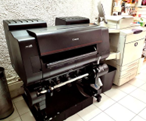 Un grand traceur d'impression noir de marque Canon est posé sur son support dans un atelier, près d'une photocopieuse blanche.