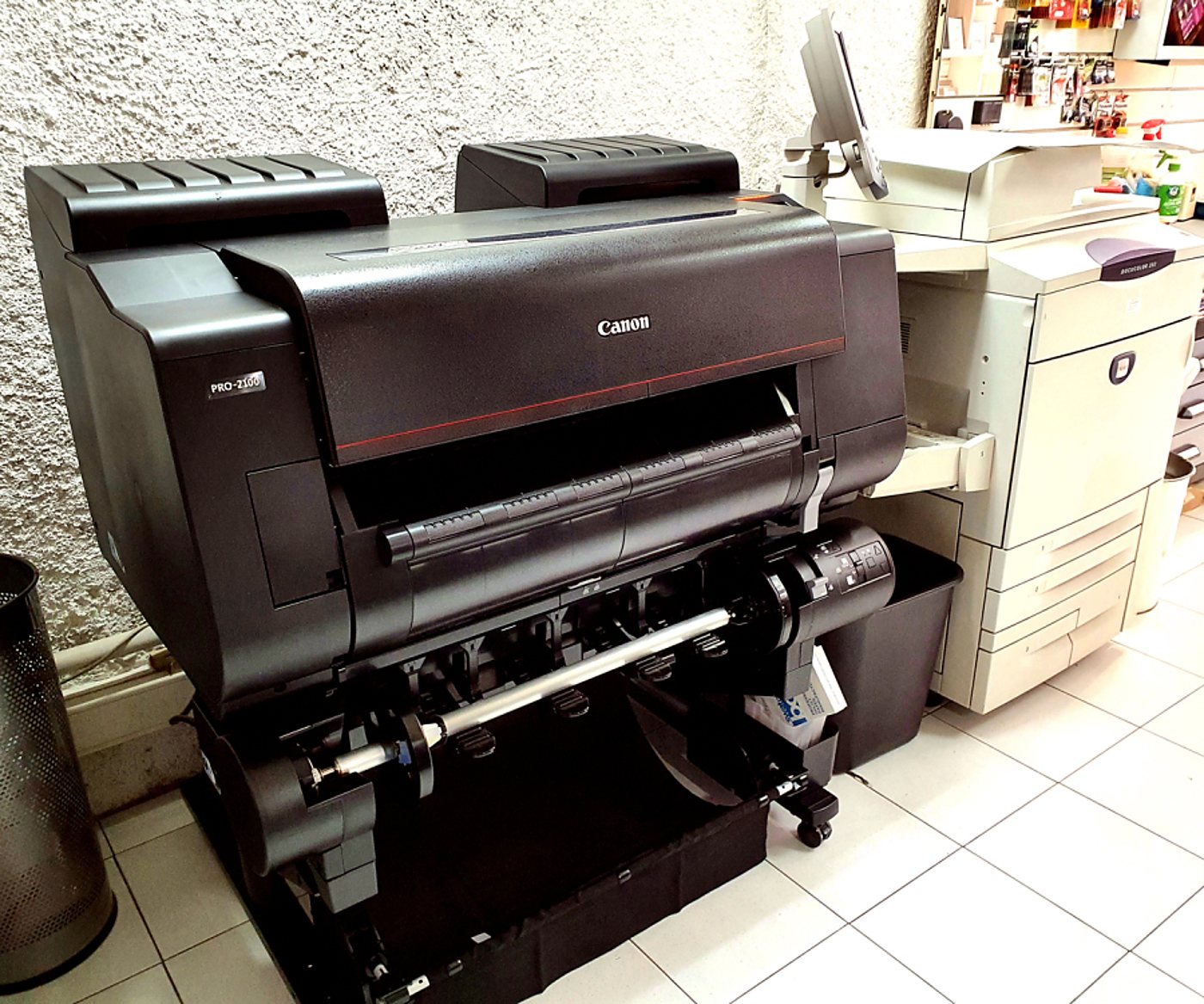 Une imprimante grand format Canon PRO-2100 noire posée sur son support, à côté d'un photocopieur et d'une corbeille, dans un local au sol carrelé.