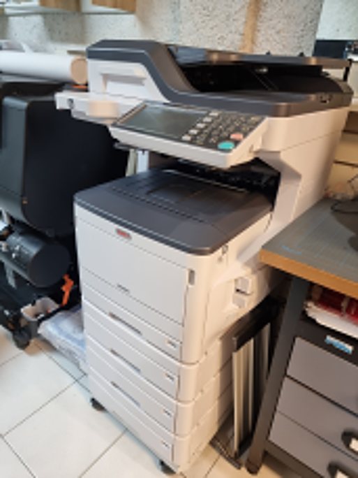 Une imprimante multifonction blanche et grise avec panneau de commande et plusieurs bacs à papier, installée dans un local encombré.