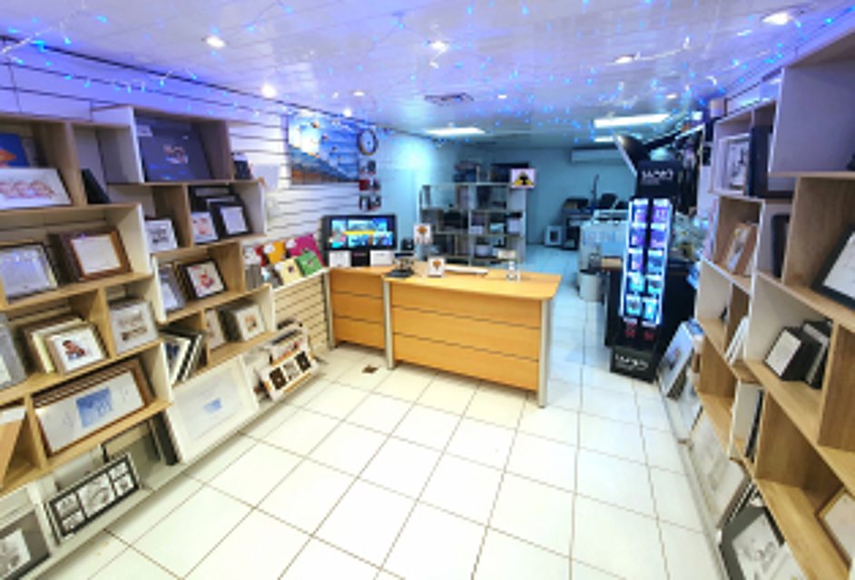 Intérieur lumineux d'une boutique présentant de nombreux cadres photo sur des étagères en bois, avec un comptoir en bois au centre et un carrelage blanc.