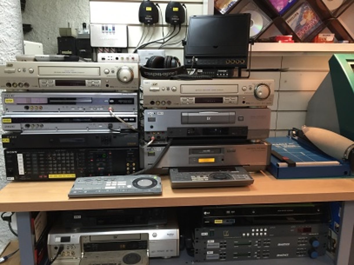 Des magnétoscopes, lecteurs DVD et autres appareils vidéo sont empilés sur une table en bois, avec deux télécommandes posées devant et d'autres appareils rangés en dessous.